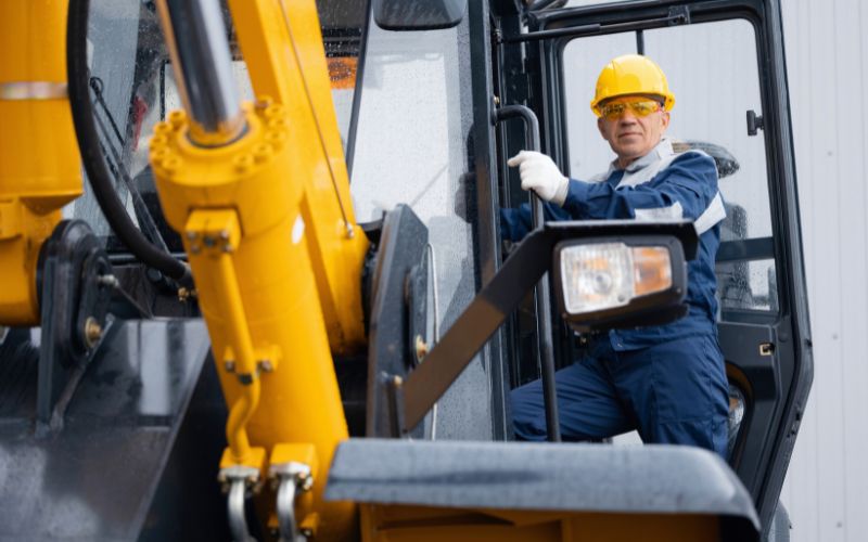 Un operaio in abiti da lavoro e casco di sicurezza giallo scende da una macchina complessa (escavatore o gru), illustrando il lavoro di operatore di macchine complesse.
