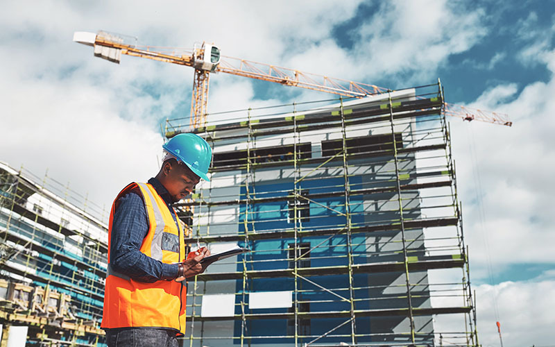 Un lavoratore edile in cantiere con casco di sicurezza e giubbotto arancione, che esamina documenti su una tavoletta digitale, con una gru e un ponteggio sullo sfondo, simboleggiando il Nuovo DL per la sicurezza sul lavoro.