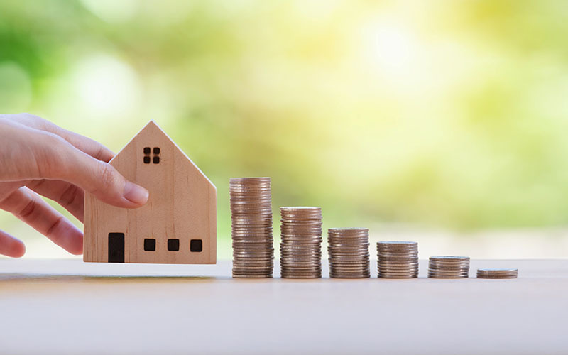 Una mano sposta una casa di legno lontano da pile di monete decrescenti, metafora della crisi abitativa e del potere d'acquisto.